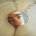 本の穴越しにこちらを見つめる人物。心の内側や夢の深層をのぞく象徴的な構図。