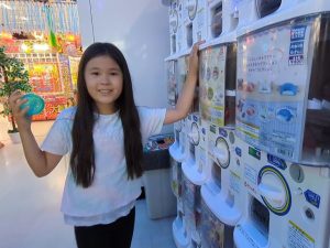 ガチャガチャの前でカプセルを手に笑顔を見せる女の子。 偶然の喜びや、小さな幸運を象徴する夢のイメージ。 A smiling girl holding a capsule toy in front of a gashapon machine, symbolizing luck, surprise, and innocent joy in dreams.