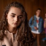 うつむく女性の後ろで、談笑するグループの姿。 悪口を言われる夢は、他人の評価を気にしすぎている心理や、自己肯定感の揺らぎを示す。 A sad young woman with people talking behind her, symbolizing fear of criticism and self-doubt.