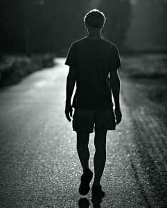 逆光の中、静かな道を一人で歩く男性の後ろ姿。ゆっくりと自分のペースに戻る象徴的なイメージ。