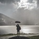 雨の湖の前で傘をさして立つ人物のモノトーン写真