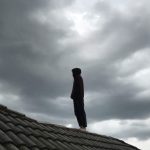 曇り空の下、家の屋根の上に立つ人物。夢の中で屋根に上がる人が示す不安定さと緊張感