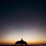 夕暮れの空の下、ベンチにひとり座る人物のシルエット
