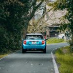木々に囲まれた細い道を進む青い車の後ろ姿。生き方の見直しや新しい道へ進む夢を象徴するイメージ
