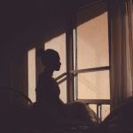 暗い部屋の窓辺に座る女性のシルエットが、外を見つめている様子